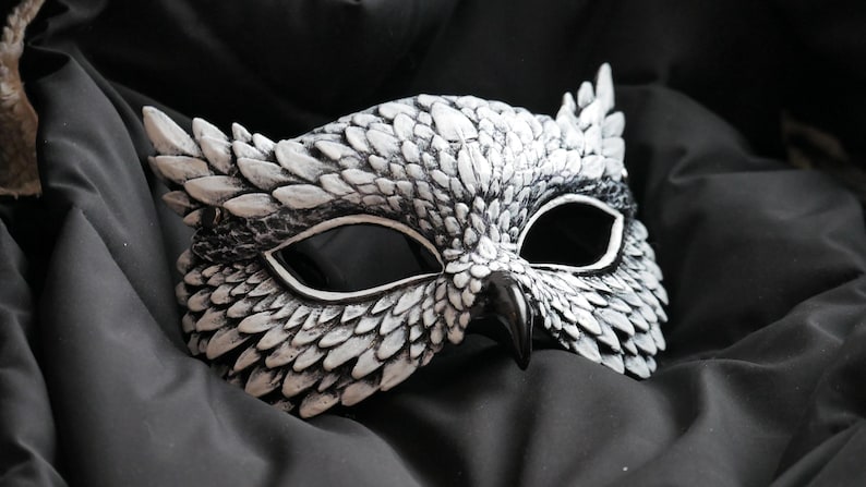 May include: A white and black owl mask with detailed feather textures. The mask has large eye openings and a prominent beak. The mask is resting on a dark, textured fabric, creating a dramatic contrast.