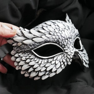 May include: A white and gray owl mask with a black strap. The mask has a detailed feather design and is held by a hand. The mask is made of a hard material and has eye openings.