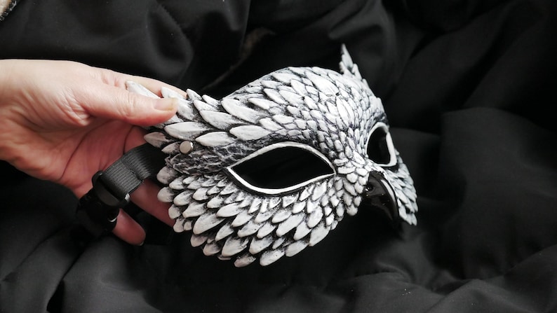 May include: A white and gray owl mask with a black strap. The mask has a detailed feather design and is held by a hand. The mask is made of a hard material and has eye openings.