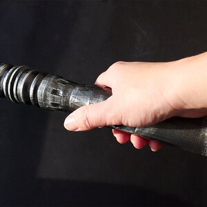 May include: A metallic silver and black lightsaber prop with detailed ridges and a slotted emitter. The prop is held in a hand against a dark background, showcasing its intricate design and sci-fi theme.