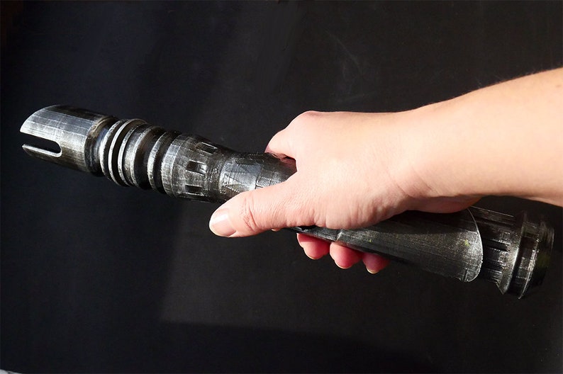May include: A metallic silver and black lightsaber prop with detailed ridges and a slotted emitter. The prop is held in a hand against a dark background, showcasing its intricate design and sci-fi theme.