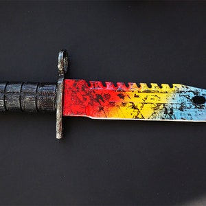 Puede incluir: Un cuchillo de colores arco&iacute;ris con mango negro. La hoja tiene un aspecto desgastado, salpicado de pintura con colores rojo, amarillo, azul y blanco.