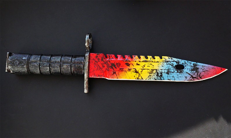 Puede incluir: Un cuchillo de colores arco&iacute;ris con mango negro. La hoja tiene un aspecto desgastado, salpicado de pintura con colores rojo, amarillo, azul y blanco.
