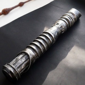 May include: A detailed, metallic silver wand with a textured, ridged design. The wand is cylindrical with a series of horizontal ridges and a textured grip. A second, brown wand is visible in the background. The wands are on a black surface.