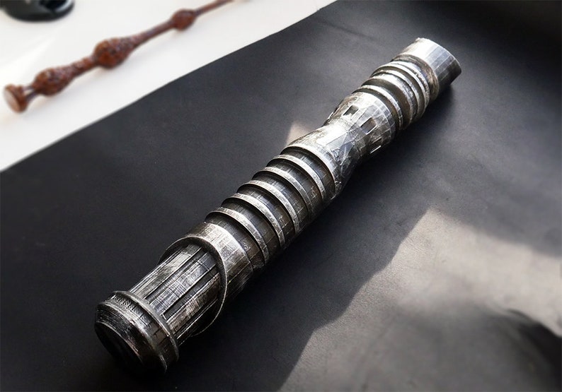 May include: A detailed, metallic silver wand with a textured, ridged design. The wand is cylindrical with a series of horizontal ridges and a textured grip. A second, brown wand is visible in the background. The wands are on a black surface.