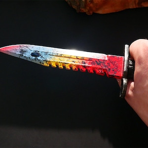 Puede incluir: Un cuchillo pintado de rojo, amarillo y azul con una hoja dentada. El cuchillo tiene un mango negro y una guarda de metal plateado.