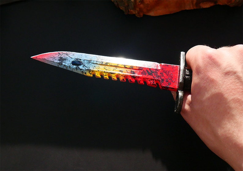 Puede incluir: Un cuchillo pintado de rojo, amarillo y azul con una hoja dentada. El cuchillo tiene un mango negro y una guarda de metal plateado.