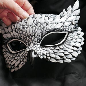 May include: A white and gray owl mask with intricate feather detailing. The mask features large eye openings and a beak, creating a realistic owl appearance. The mask is held against a dark background, highlighting its design.
