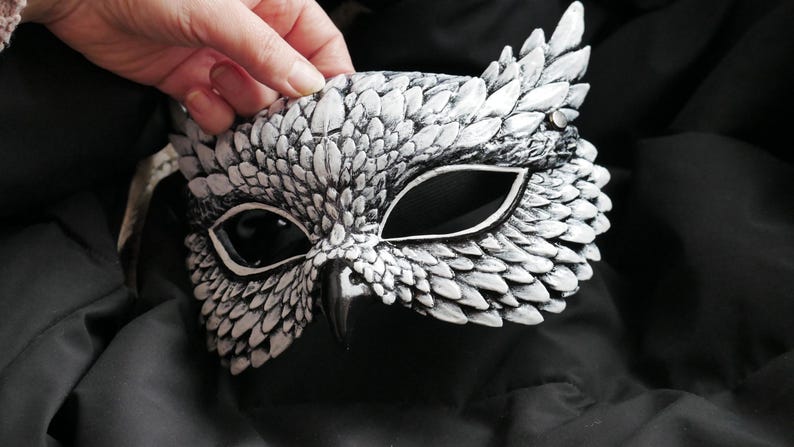 May include: A white and gray owl mask with intricate feather detailing. The mask features large eye openings and a beak, creating a realistic owl appearance. The mask is held against a dark background, highlighting its design.