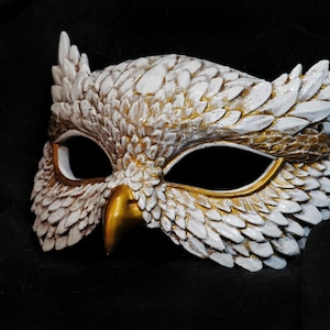 May include: A white and gold owl mask with intricate feather detailing. The mask features a gold beak and eye accents, and is set against a dark background. This decorative mask is suitable for costume parties or theatrical performances.