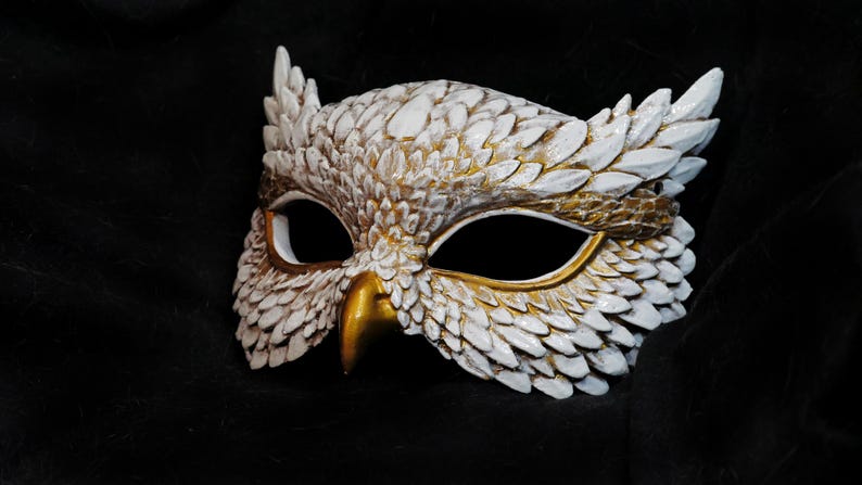 May include: A white and gold owl mask with intricate feather detailing. The mask features a gold beak and eye accents, and is set against a dark background. This decorative mask is suitable for costume parties or theatrical performances.