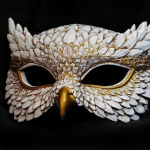 May include: A handcrafted owl mask with a white and gold color scheme. The mask features detailed feather-like textures, a gold beak, and large eye openings. The mask is designed for costume or decorative purposes.