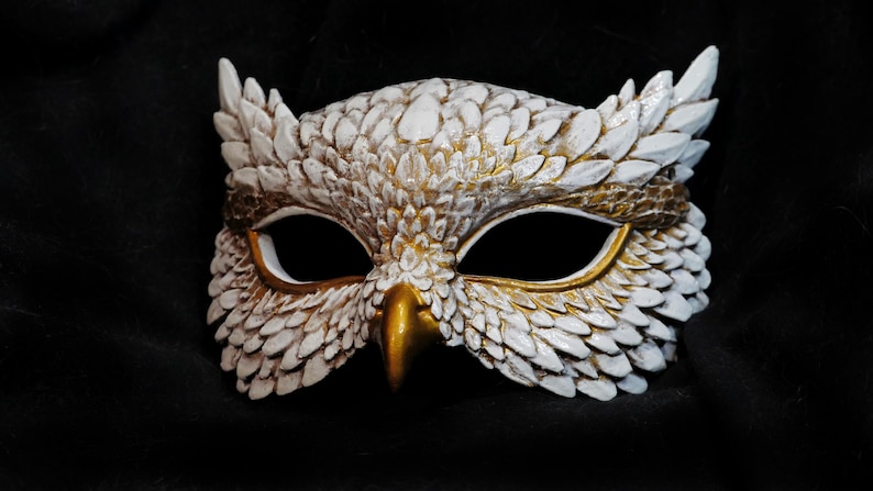 May include: A handcrafted owl mask with a white and gold color scheme. The mask features detailed feather-like textures, a gold beak, and large eye openings. The mask is designed for costume or decorative purposes.