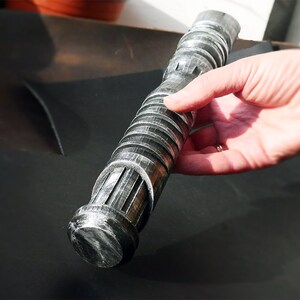 May include: A silver and black lightsaber prop with a ribbed handle and detailed emitter. The prop is held by a hand, showcasing its cylindrical design and metallic finish. The lightsaber is a replica of a popular science fiction weapon.