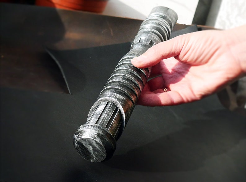 May include: A silver and black lightsaber prop with a ribbed handle and detailed emitter. The prop is held by a hand, showcasing its cylindrical design and metallic finish. The lightsaber is a replica of a popular science fiction weapon.