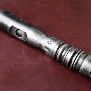 May include: A metallic silver lightsaber prop with a detailed, textured design. The prop features a cylindrical body with various ridges, grooves, and a beveled end. The lightsaber is displayed on a dark red surface.