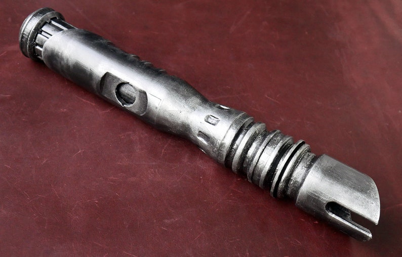 May include: A metallic silver lightsaber prop with a detailed, textured design. The prop features a cylindrical body with various ridges, grooves, and a beveled end. The lightsaber is displayed on a dark red surface.