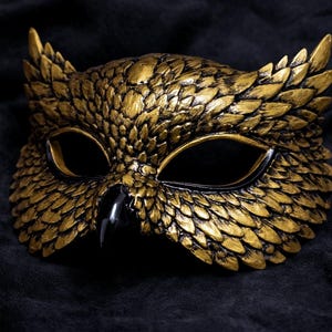 May include: A gold-colored owl mask with black eye openings and beak. The mask has a textured, feathered appearance and is set against a dark background, creating a dramatic effect. Ideal for costume parties.