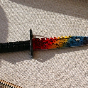 Puede incluir: Un cuchillo de colores arco&iacute;ris con mango negro. La hoja tiene un borde dentado y un patr&oacute;n de arco&iacute;ris con detalles negros.