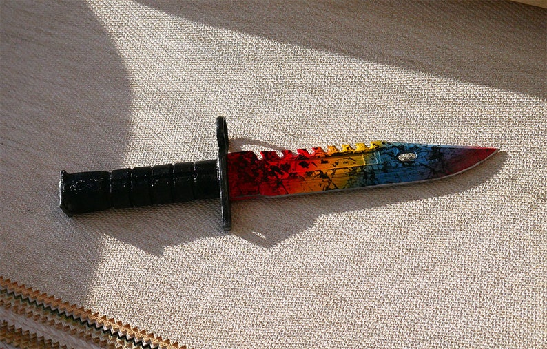 Puede incluir: Un cuchillo de colores arco&iacute;ris con mango negro. La hoja tiene un borde dentado y un patr&oacute;n de arco&iacute;ris con detalles negros.