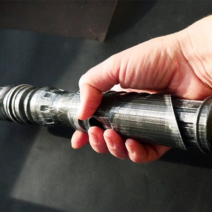 May include: A metallic gray lightsaber hilt is held in a hand. The hilt has a cylindrical design with detailed grooves and ridges. The overall aesthetic suggests a futuristic or science fiction theme.