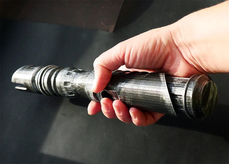 May include: A metallic gray lightsaber hilt is held in a hand. The hilt has a cylindrical design with detailed grooves and ridges. The overall aesthetic suggests a futuristic or science fiction theme.