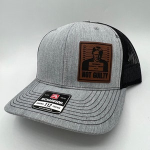 May include: Gray and black trucker hat with a Richardson patch. The hat features a brown leather patch with an image of a person in a mugshot and the words "NOT GUILTY". The hat is adjustable.