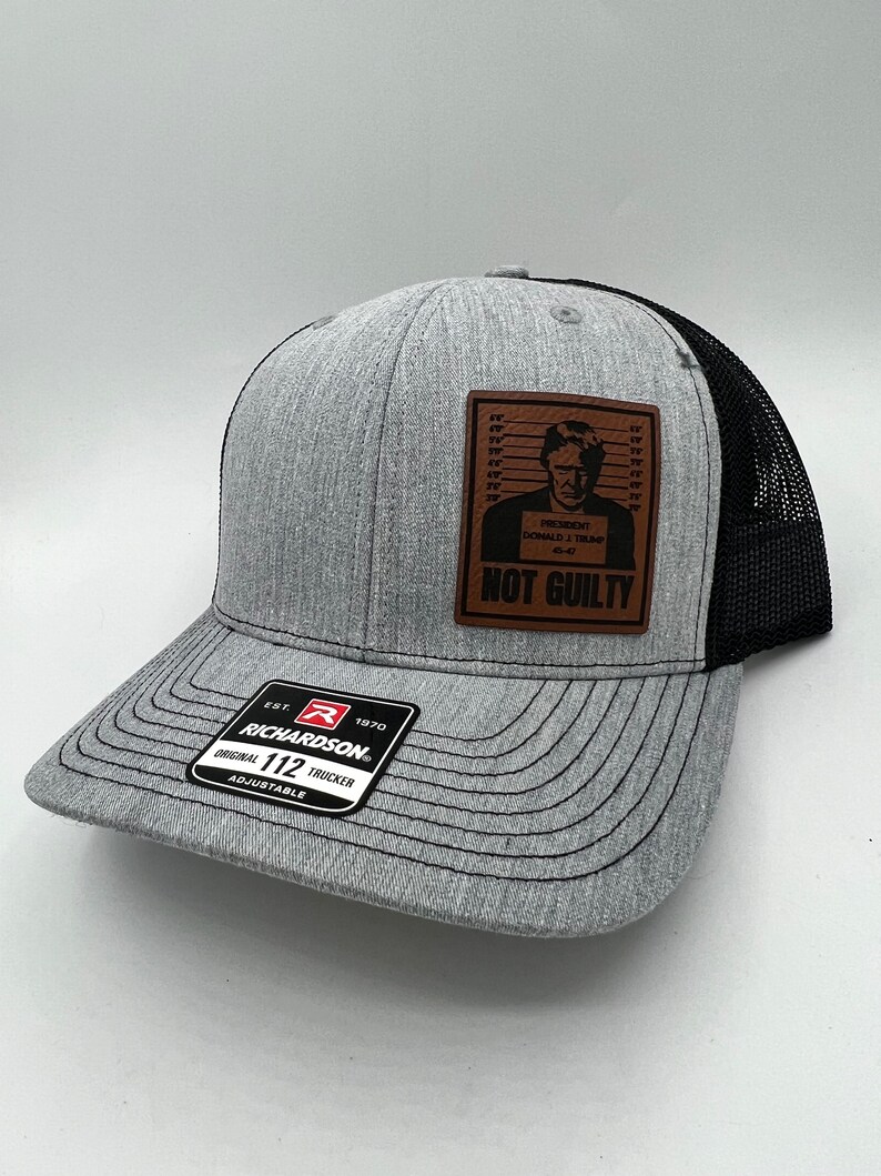 May include: Gray and black trucker hat with a Richardson patch. The hat features a brown leather patch with an image of a person in a mugshot and the words "NOT GUILTY". The hat is adjustable.
