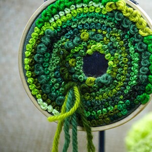 May include: A circular embroidery hoop with a green and white yarn design. The yarn is arranged in a spiral pattern, creating a circular shape in the center. The yarn is a variety of shades of green, from light to dark.