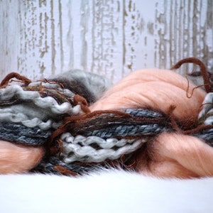 May include: A pile of soft, fluffy yarn in shades of peach, gray, and brown. The yarn is arranged in a loose, flowing pile, with some strands of yarn extending out from the pile.