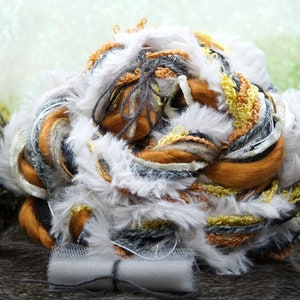 May include: A ball of yarn with a mix of white, gray, brown, and yellow fibers. The yarn is fluffy and textured, with some strands of metallic thread. The yarn is sitting on a brown surface.