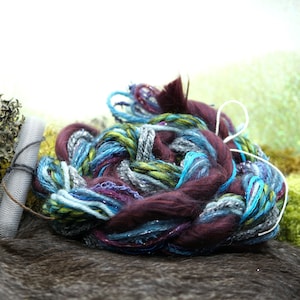 May include: A close-up of a ball of yarn with a mix of purple, blue, green, and gray strands. The yarn is soft and fluffy, and it has a subtle shimmer.