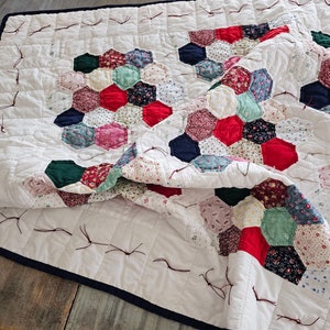Peut inclure: Une courtepointe blanche avec une bordure bleu marine. La courtepointe est faite de tissu patchwork de différentes couleurs et motifs, notamment rouge, vert et rose. La courtepointe est pliée en deux et a quelques fils lâches qui pendent des bords.