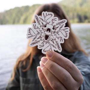 Puede incluir: Una pegatina blanca con una ilustración marrón de una calavera rodeada de huesos. La pegatina se sostiene frente a una persona, con un fondo borroso de un lago y árboles.