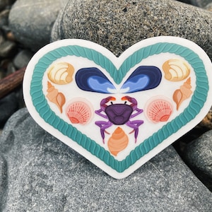 May include: A heart-shaped sticker featuring a teal rope border. Inside, various seashells, a purple crab, and blue mussels are arranged. The sticker is set against a background of gray rocks.