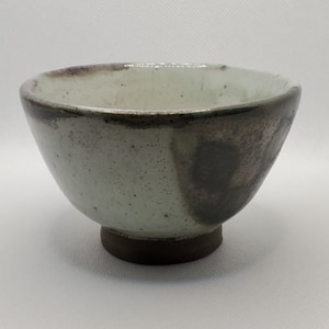 Puede incluir: Un pequeño cuenco de cerámica blanca con un borde marrón y una base marrón. El cuenco tiene un esmalte moteado y una mancha marrón oscura en el lateral.