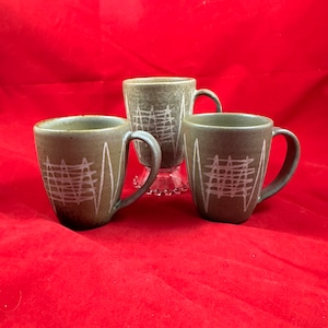 May include: Three green ceramic mugs with white geometric designs. The mugs have a rounded shape and a handle on the side.