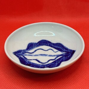 Puede incluir: Un tazón de cerámica blanca con un diseño de labios azul y blanco. El tazón está sobre una superficie roja.