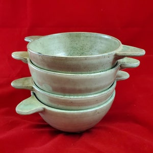 Puede incluir: Un conjunto de cuatro cuencos de cerámica verde con asas. Los cuencos están apilados uno encima del otro, con el cuenco más grande en la parte inferior y el más pequeño en la parte superior.