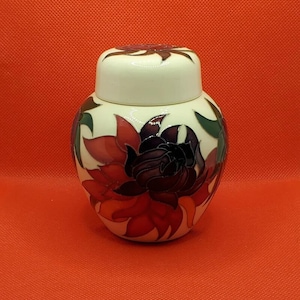 Może przedstawiać: Biały ceramiczny dzbanek z kwiatowym wzorem. Dzbanek ma pokrywkę i jest ozdobiony czerwonymi, fioletowymi i zielonymi kwiatami.