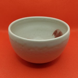 Könnte beinhalten: Eine weiße Keramikschüssel mit einem kleinen roten Design auf der Innenseite. Die Schüssel ist rund und hat einen leicht ausgestellten Rand.