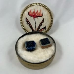 Puede incluir: Dos pequeñas baldosas cuadradas de cerámica con un esmalte azul y un diseño blanco. Las baldosas están en una caja redonda blanca con un diseño floral en la tapa.