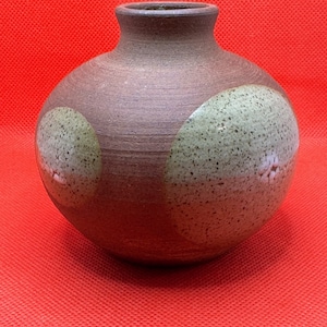 Puede incluir: Un jarrón de cerámica marrón con dos círculos grandes de color blanco roto con motas verdes. El jarrón tiene un cuerpo redondeado y un cuello estrecho.