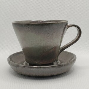 Puede incluir: Un juego de taza y platillo de cerámica gris con un sutil glaseado verde y marrón. La taza tiene una forma ancha y redondeada y un asa. El platillo es ligeramente más grande que la taza y tiene un glaseado a juego.