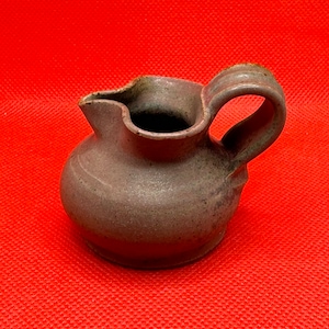 Puede incluir: Una pequeña jarra de cerámica gris con cuerpo redondeado y un labio ligeramente acampanado. La jarra tiene una sola asa y un esmalte moteado.