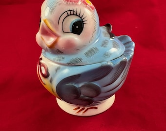 May include: A blue ceramic bird with a pink beak and large eyes. The bird has a flower on its head and is decorated with blue and white stripes. The bird is a sugar bowl with a lid.