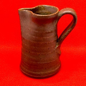 Puede incluir: Una jarra de cerámica marrón hecha a mano con asa y pico vertedor. La jarra tiene un aspecto rústico y texturizado y está sobre un fondo rojo. La jarra mide aproximadamente 15 cm de altura.