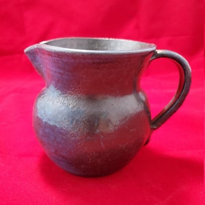 Puede incluir: Una jarra de cerámica oscura, hecha a mano, con un cuerpo redondeado y un asa curvada. La jarra tiene un pico para verter fácilmente y un esmalte oscuro y texturizado con toques de azul. El fondo es rojo liso.