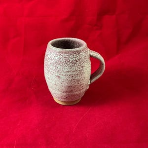 Puede incluir: Una taza de cerámica blanca con un esmalte morado moteado y un asa. La taza tiene un cuerpo redondeado y un borde ligeramente acampanado.