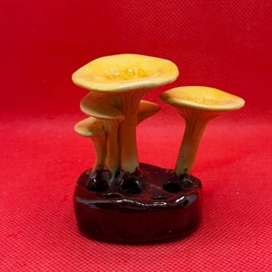 May include: A ceramic sculpture featuring a cluster of four yellow-capped mushrooms with slender stems emerging from a dark brown, glossy, rounded base. The mushroom caps have a smooth, concave surface, and the stems are attached to the dark base.